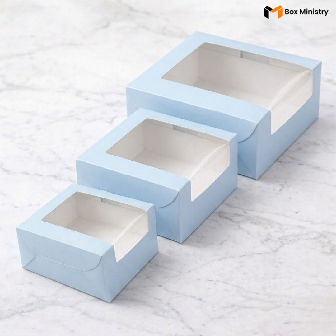 Three pastel blue cake boxes in three sizes with L-shape PVC windows arranged on white marble surfac