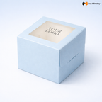 Soft sky blue square cake box with large square clear PVC window on lid showing "YOUR LOGO" text in elegant serif grey font centered on white interior base, positioned at 3/4 isometric angle, tall upright shape with prominently visible side walls, visible tuck-lock flap on right side face, matte finish, on a soft pale blue to white gradient background