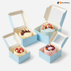 Four open sky blue square window cake boxes arranged in a 2x2 grid composition, each containing a uniquely decorated cake — ivory cake with berries and gold leaf, sky blue frosted cake with pink roses and macarons, white cream cake with strawberries and pink macarons, and pink frosted cake with blueberries and edible flowers — all lids open showing clear PVC windows and white interior lining, on a soft pure white background