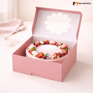 Open pink flower window cake box with lid propped upright showing decorative flower-shaped PVC window, filled with a round blush pink frosted celebration cake decorated with fresh strawberries, edible purple and white flowers, and green leaves — Pink Flower Window Cake Box .