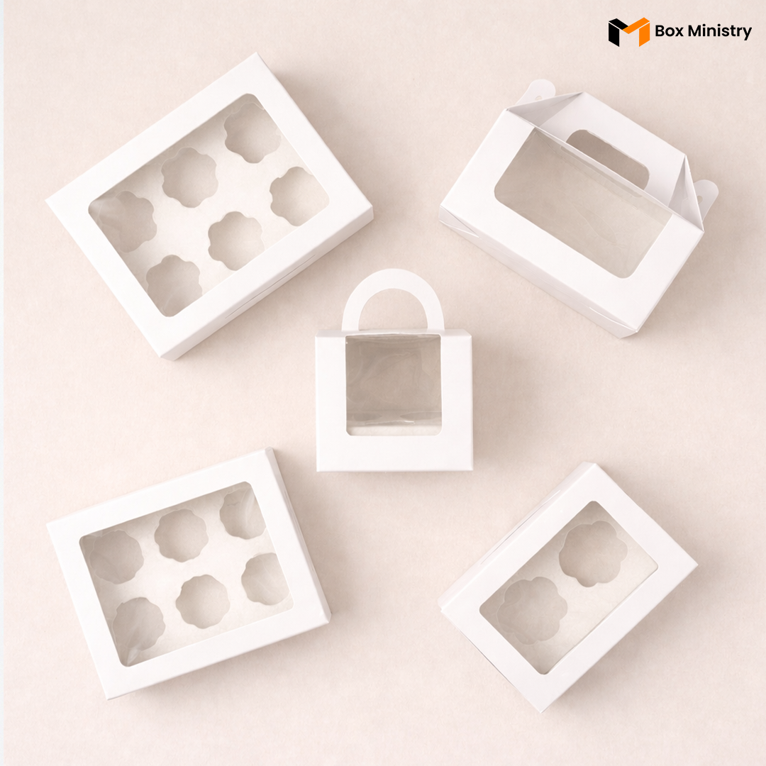 White cupcake boxes in multiple sizes with window lids and gable handles, arranged in a flat lay product display

